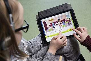 Elev med hodetelefoner som bruker et nettbrett som viser « Friends »-appen med en illustrert scene og en «Tester»-knapp