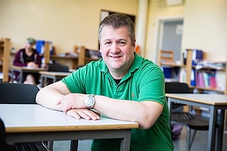Smilende mann i en grønn poloskjorte sitter ved et skrivebord i et klasserom