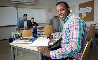 Smilende mann i rutete skjorte skriver i en notatbok ved et skrivebord i et klasserom, to personer jobber ved datamaskiner bak ham