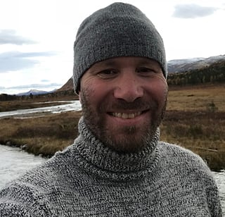 Smilende skjeggete mann i grå lue og strikket genser utendørs ved en elv i fjellterreng