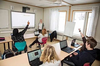 Elever som rekker opp hendene i et klasserom mens en lærer står ved en storskjerm